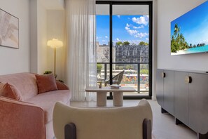 A modern living room with a pink sofa, a table with a book, a lamp, a TV on the wall, and a view of buildings and a beach. - Nomad condo (Miami)