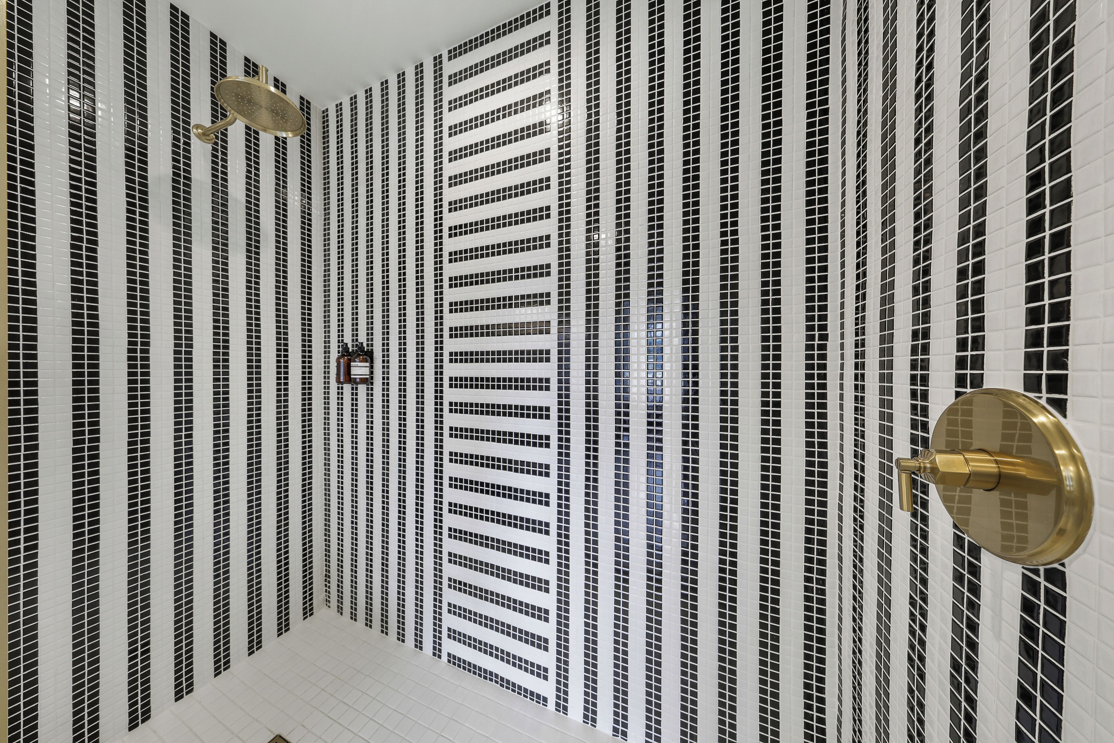 Casa de banho com parede revestida em azulejos, apresentando um padrão vertical em cores preto e branco, uma torneira dourada e um chuveiro dourado.