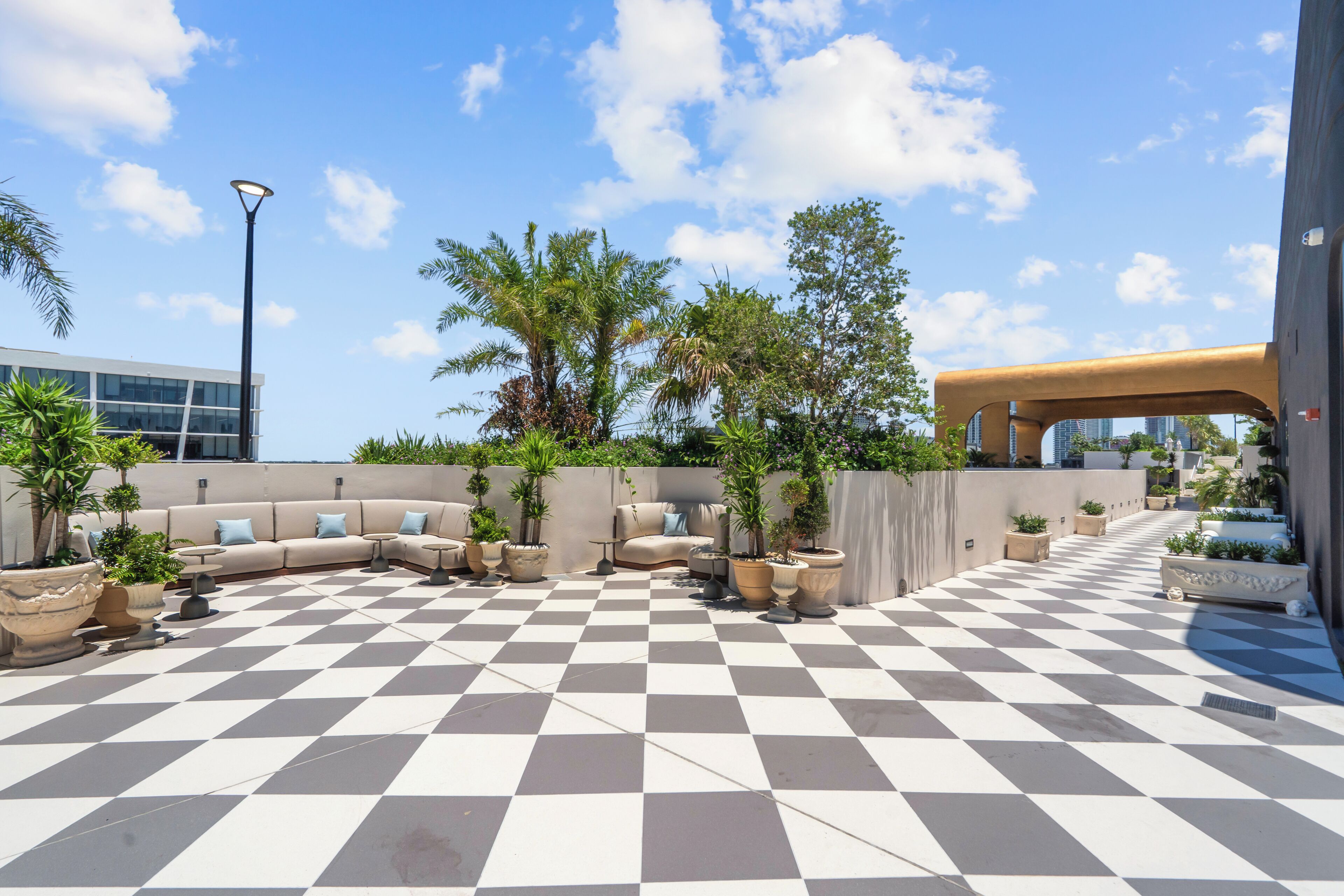A rooftop terrace with a checkered floor, outdoor seating, potted plants, and a modern building in the background.
