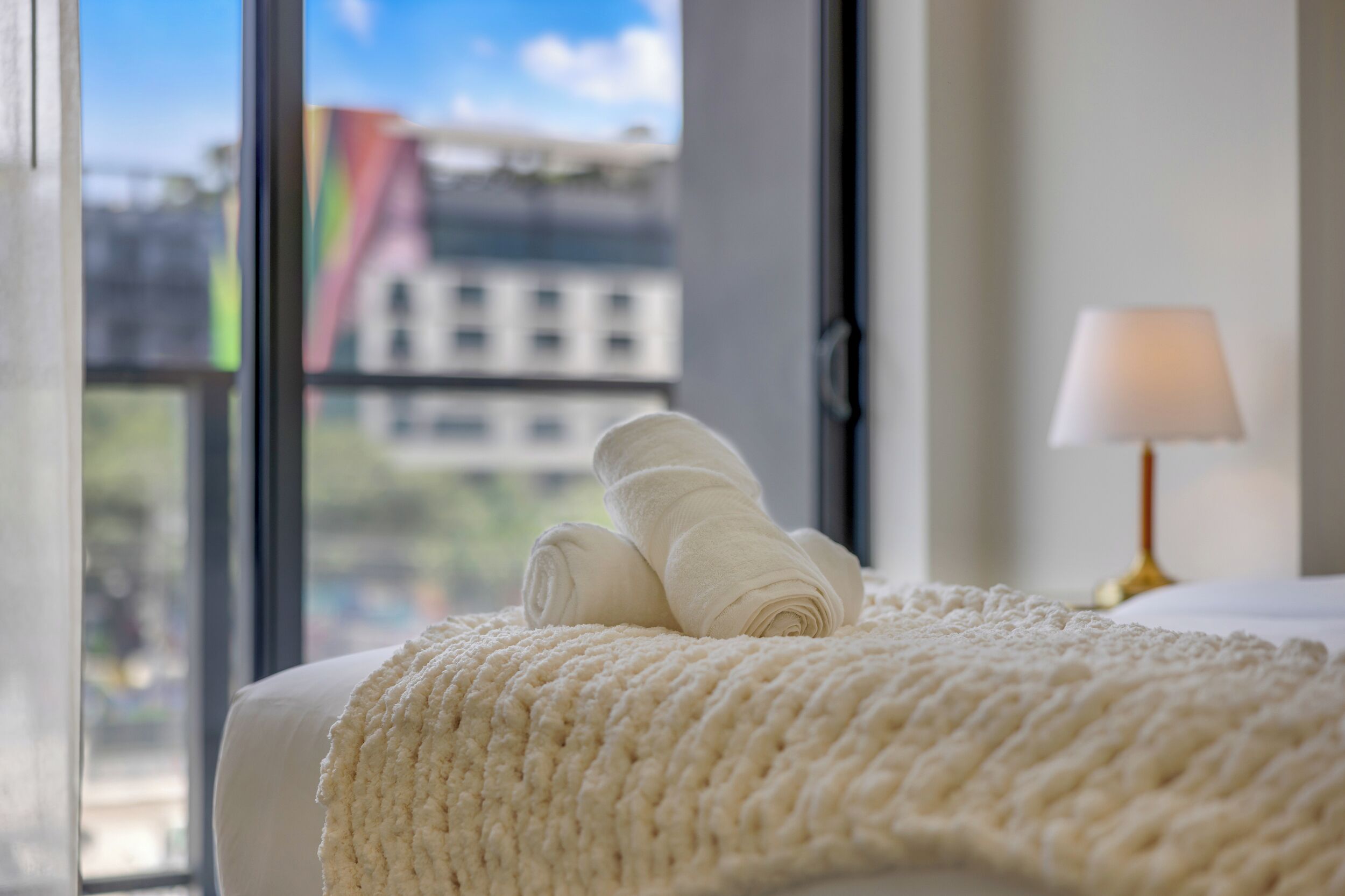 A bed with a white blanket and rolled towels, a lamp, and a view of buildings through a window.