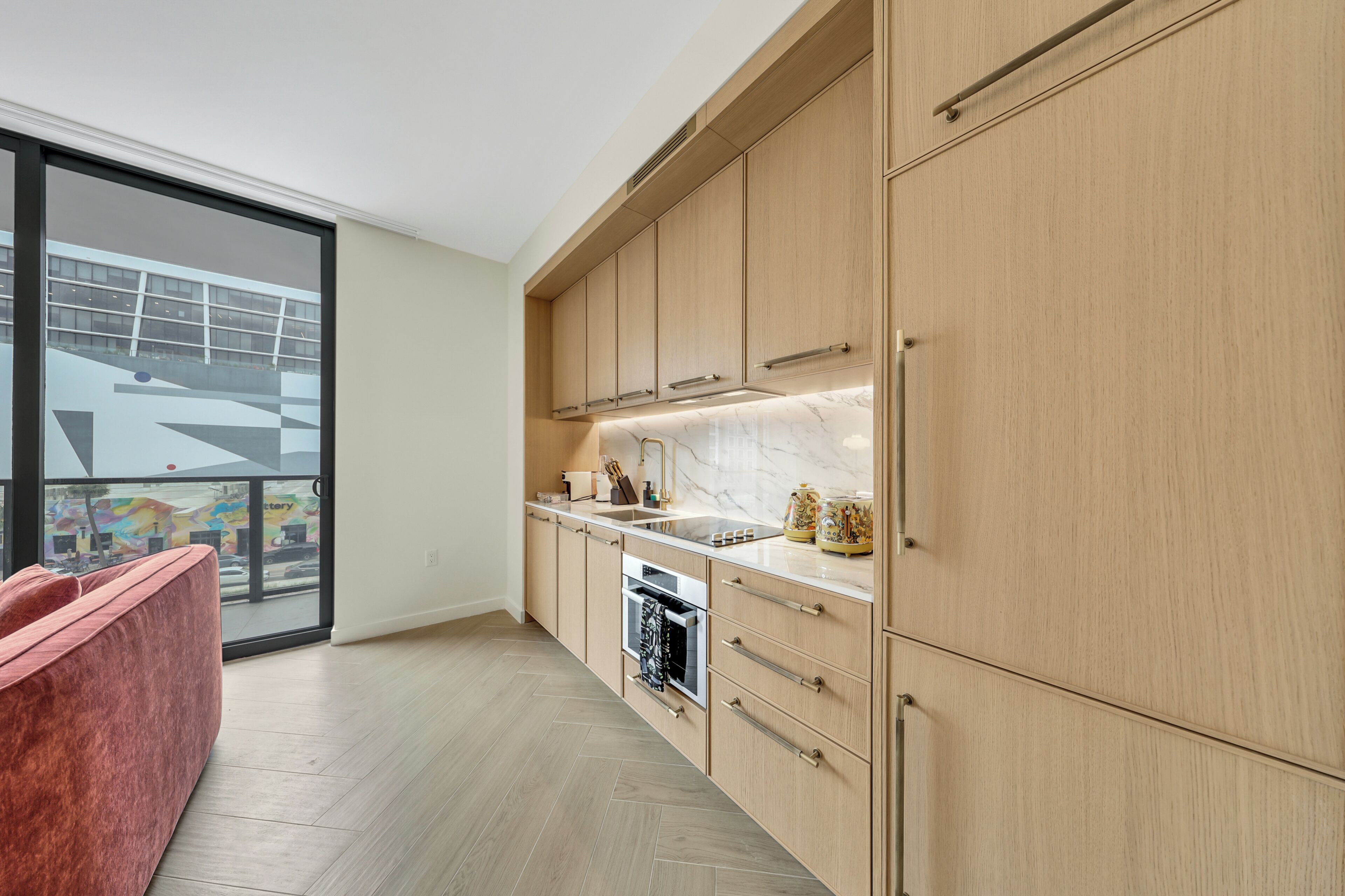 Eine moderne Küche mit Holzschränken, einem eingebauten Ofen und einem großen Fenster mit Blick auf einen farbenfrohen Außenbereich.