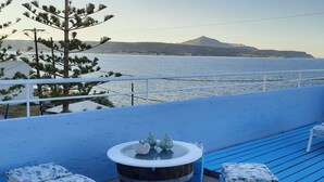 Una terraza frente al mar con mobiliario azul, una mesa redonda con un objeto decorativo y vistas al mar y colinas distantes.