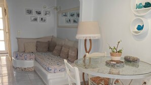 Una sala de estar con una mesa de vidrio, sillas blancas, un sofá con cojines estampados, una lámpara y estantes montados en la pared con objetos decorativos.