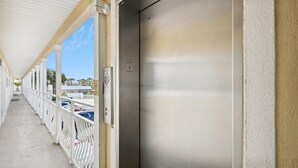 A covered corridor with a metal elevator, white railings, and a view of parked cars and buildings. - Crystal Sands 215a- Saltwater Daze (Destin)