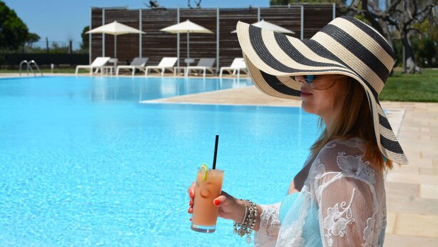 A poolside scene with a person holding a drink, lounge chairs, and umbrellas in the background. - Masseria Corda di Lana - Suite Deluxe (Leverano)
