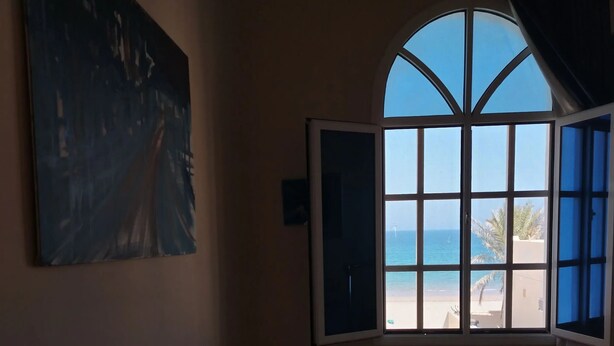 Une pièce avec une grande fenêtre en arc de cercle, un bureau, une chaise, un tableau accroché au mur et une petite plante.
