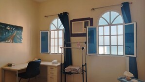 Une pièce comprenant un bureau, une chaise, un lit, deux fenêtres avec des rideaux, un radiateur et une petite table agrémentée d’objets décoratifs.