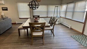 Un comedor con mesa y sillas de madera, una araña y ventanas amplias con persianas.