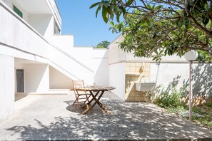 Um pátio com mesa e cadeiras, um edifício branco e uma árvore.
