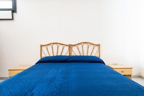 Sängyssä on sininen peitto, puinen pääty ja pieni puinen yöpöytä, jossa on lamppu.