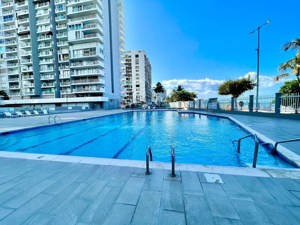 A swimming pool with clear blue water, surrounded by a tiled deck and a tall residential building in the background.