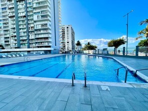 A swimming pool with clear blue water, surrounded by a tiled deck and a tall residential building in the background. - Isla Verde Beach/Self Check-in, 1 Parking, Pool (Carolina)
