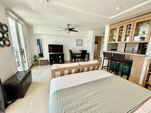 A modern bedroom with a bed, a ceiling fan, a TV, a dining area, and a kitchen with wooden cabinets. - Isla Verde Beach/Self Check-in, 1 Parking, Pool (Carolina)