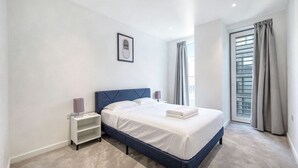 A bedroom with a bed, a nightstand, a wall-mounted picture, and a window with curtains. - Air conditioner 2 Bedroom central flat (London)