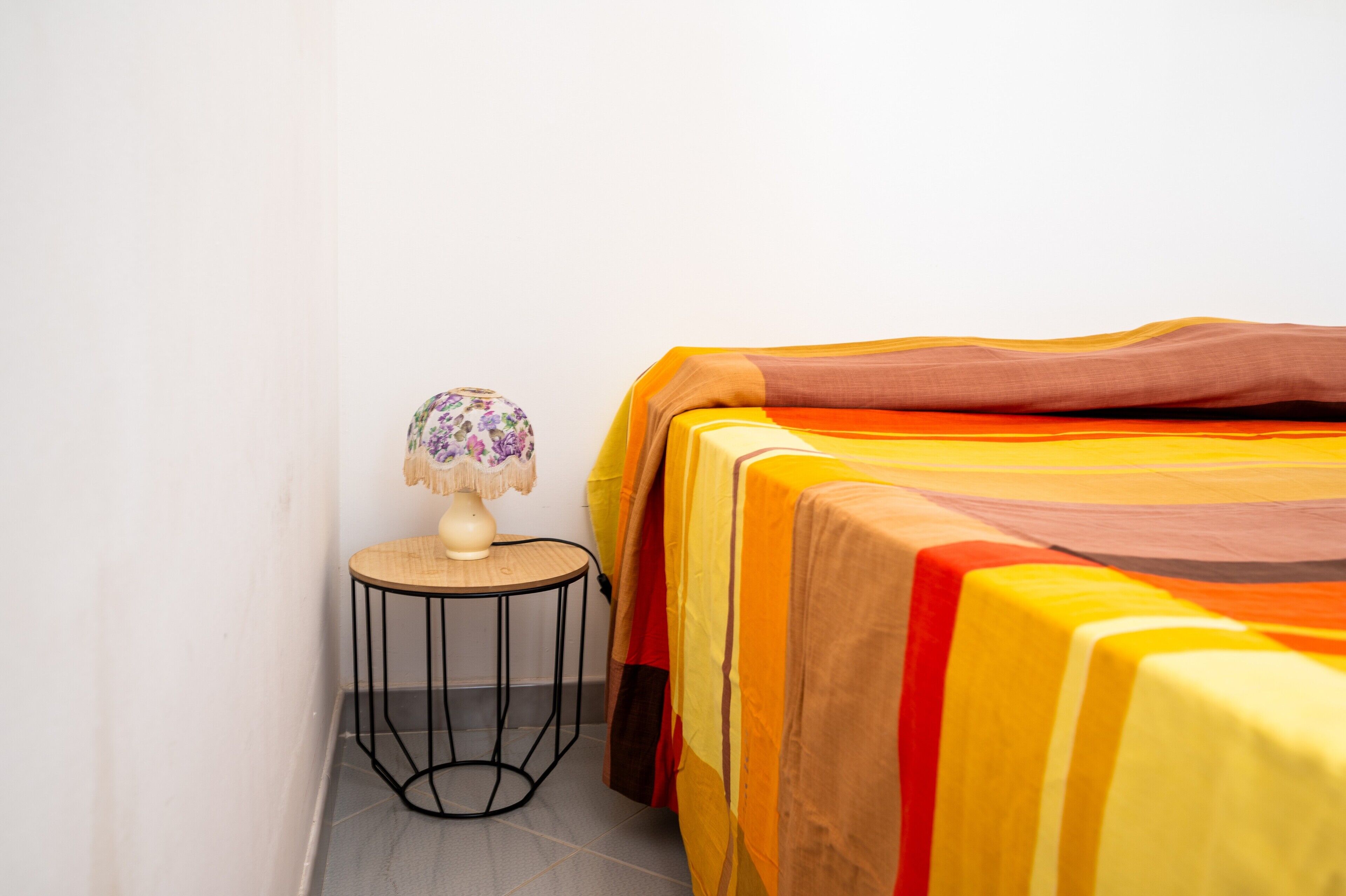 A bed with a colorful blanket, a small round table with a lamp, and a plain wall.