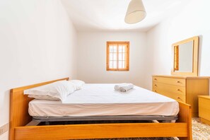 Ein ordentlich bezogenes Bett mit weißen Leinen, ein hölzernes Kopfteil, ein kleines Fenster mit Holzrahmen, eine Holzkommode und ein Spiegel an der Wand.