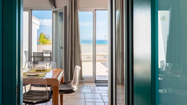 A room with a dining table set for two, a chair, and a view of the sea through a glass door. - Apartment 'Terrazza Sul Mare' with Sea View, Private Terrace and Air Conditioning (San Foca)