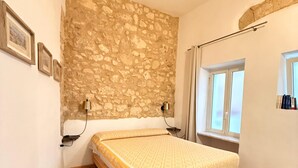 Quarto com parede de pedra, piso de madeira, cama, mesa de cabeceira, luminária e janela.