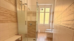 Casa de banho com chuveiro, sanita e lavatório, chão em madeira e um banco.