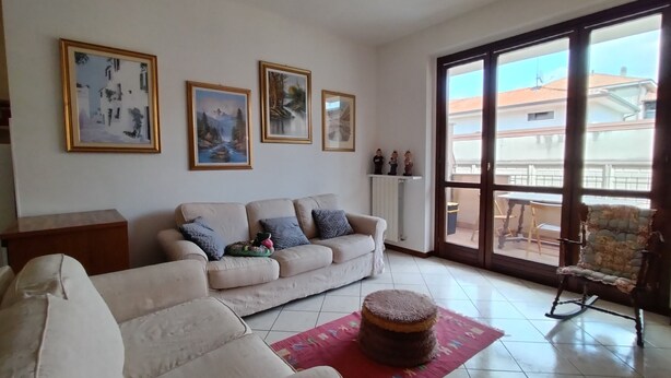Ein Wohnzimmer mit einer Couch, einem Couchtisch und einem Schaukelstuhl. An den Wänden hängen gerahmte Bilder und ein Fenster bietet Blick auf Gebäude.