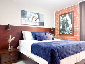Ein Hotelzimmer mit Bett, Nachttisch, einer Blumenvase, einer Wandlampe und einem gerahmten Bild.