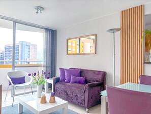 Ein modernes Wohnzimmer mit einem violetten Sofa, einem weißen Sessel, einem Glastisch und Blick auf Gebäude durch große Fenster.