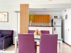 Ein modernes Wohnzimmer mit einem violetten Sofa, einem Esstisch mit violetten Stühlen, einer Küchenzeile mit Holzschränken und einer Mikrowelle.