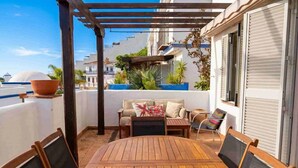 A wooden table and chairs under a pergola, with a sofa and cushions, and a view of buildings in the background. - Oasis Quitapenas Apartment Arroyo Marinos with Pool, Shared Terrace & Wi-Fi (Almuñécar)