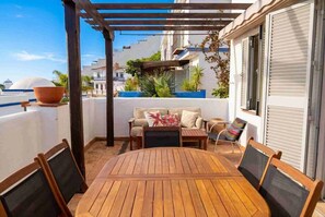 Ett träbord och stolar under en pergola, med en soffa och kuddar, och en utsikt över byggnader i bakgrunden.