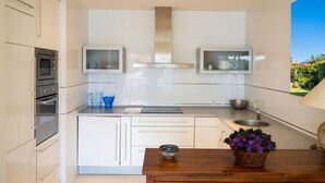 A modern kitchen with white cabinetry, a built-in oven, a stainless steel range hood, and a window overlooking a garden. - Oasis Quitapenas Apartment Arroyo Marinos with Pool, Shared Terrace & Wi-Fi (Almuñécar)