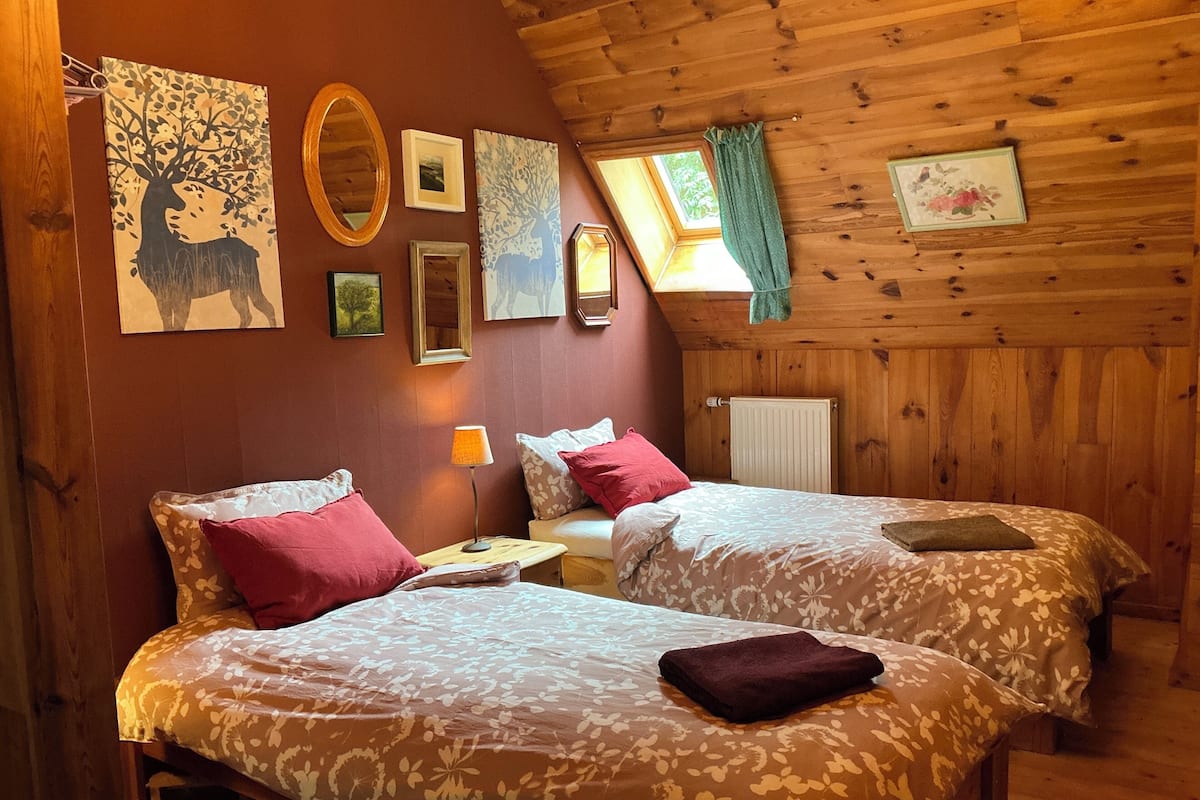 A cozy room with wooden walls, two beds with floral bedding, a nightstand with a lamp, and artwork on the walls.