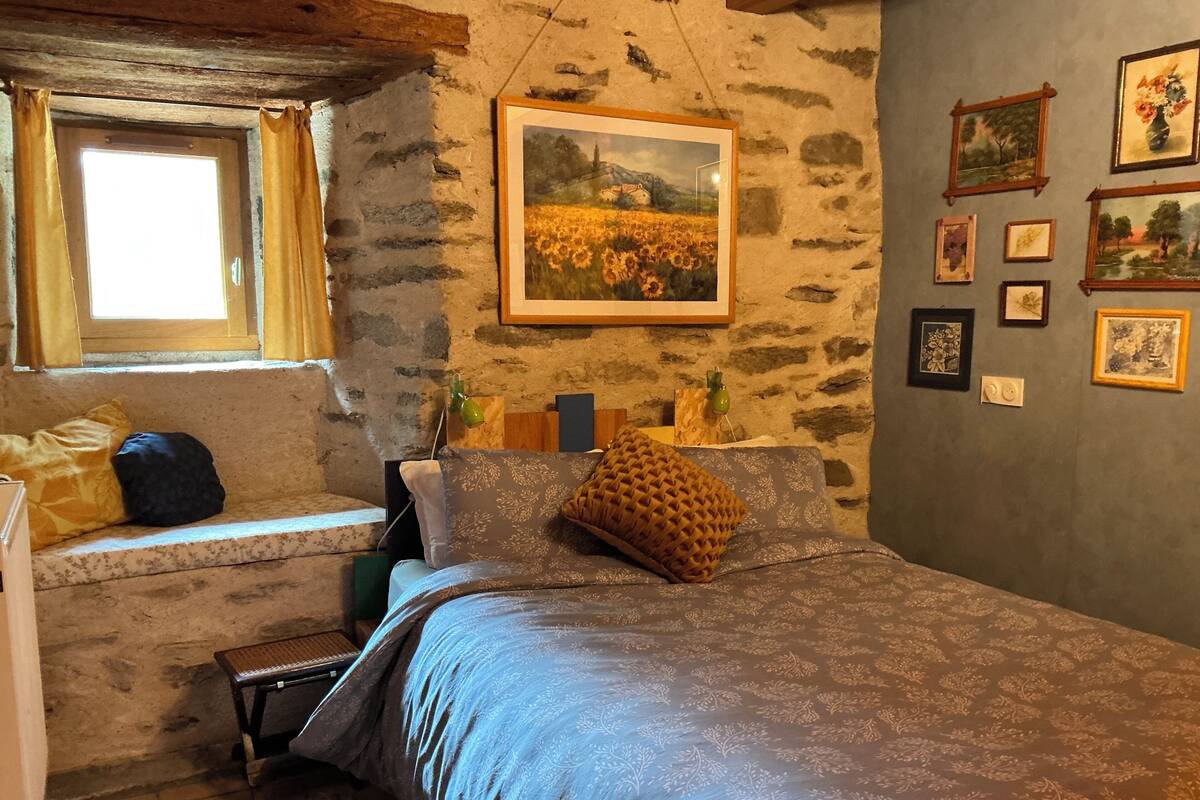 A bedroom with a stone wall, a bed with patterned bedding, a bench by the window, and framed pictures on the wall.