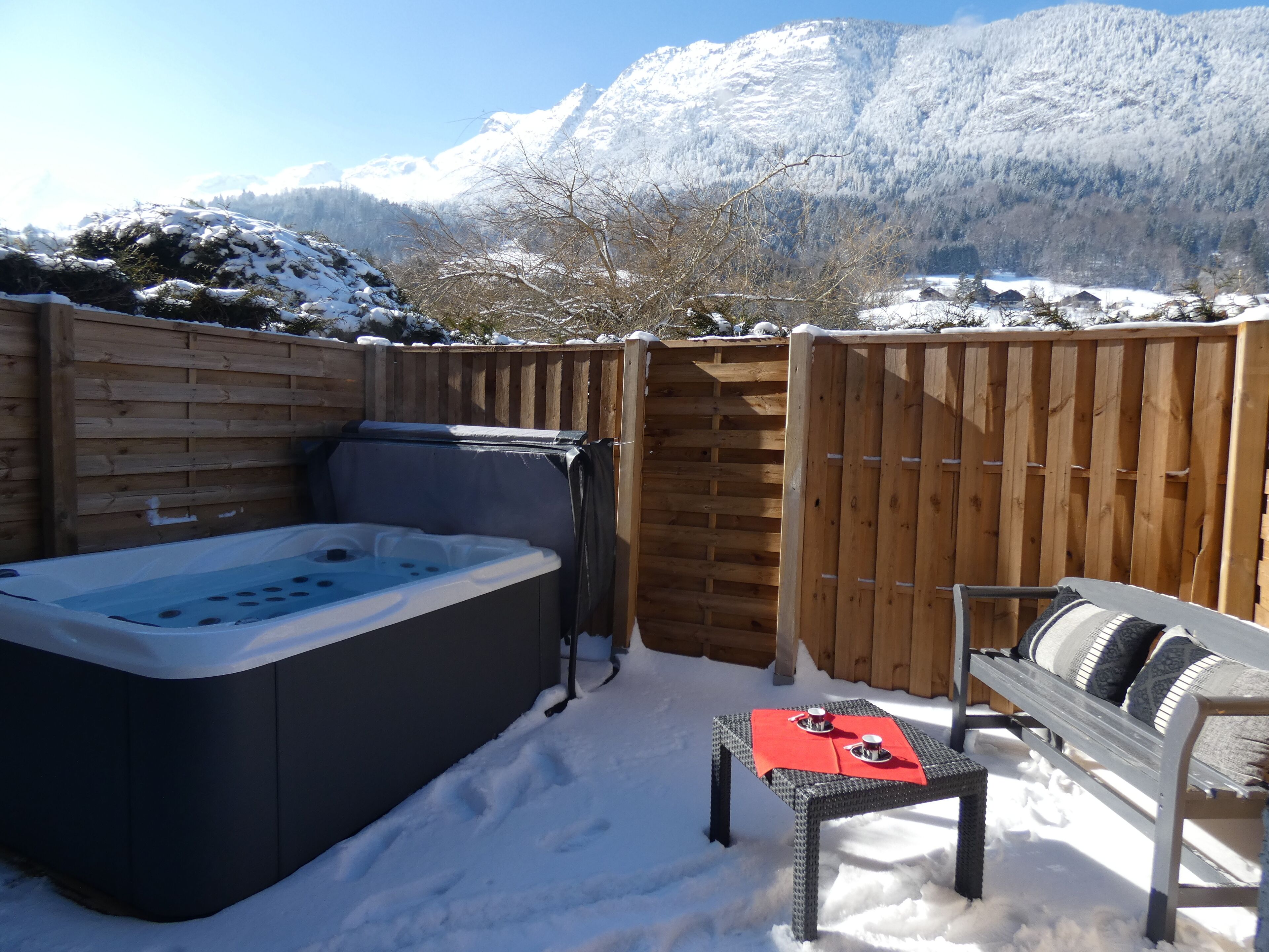 A hot tub, a bench, and a table with red placemats in a snowy outdoor area with a wooden fence and mountains in the background.