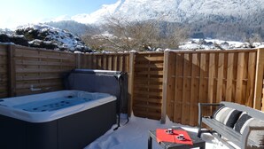 A hot tub, a bench, and a table with red placemats in a snowy outdoor area with a wooden fence and mountains in the background. - Apartment DAHU Domaine du Fraizier Thônes private jacuzzi annecy and shops (Thônes)