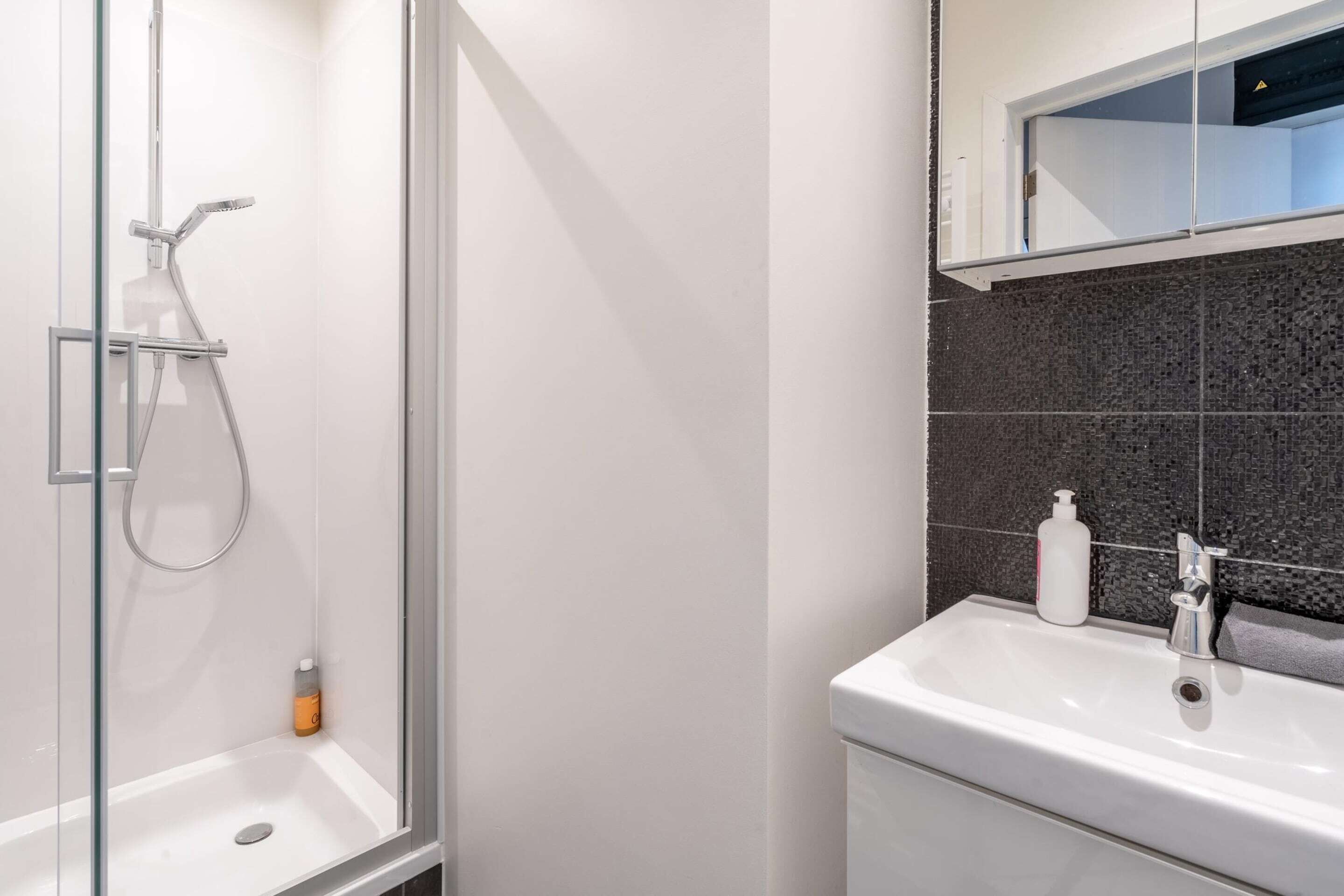 Un baño moderno con ducha envuelta en vidrio, un lavamanos blanco y una pared revestida de baldosas oscuras.