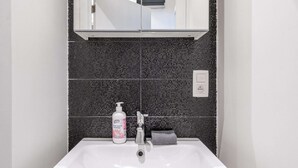 Un baño con un lavabo blanco, un salpicadero de azulejos oscuros, un espejo sobre el lavabo y una botella de jabón para manos.