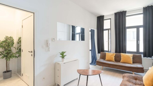Una sala de estar moderna con un sofá, una mesita, una planta en maceta, un mueble blanco, un espejo y una ventana con cortinas.
