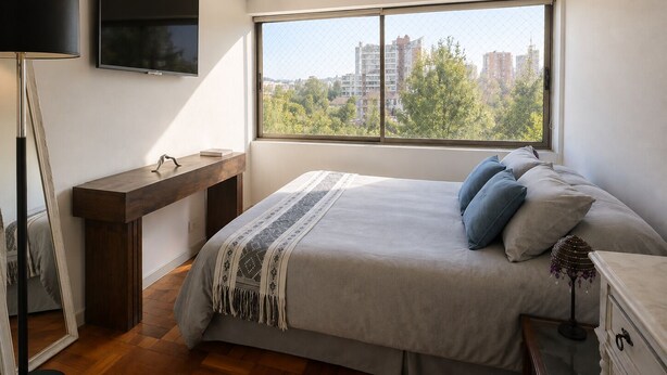 A bedroom with a large window, a bed with pillows, a bedside table, a TV mounted on the wall, and a floor lamp. - Luminous Home Studio with privileged view to the Park. Steps away from Metro Bilbao. Wifi (Providencia)