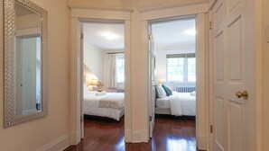 Een gang die leidt naar twee slaapkamers, met houten vloeren, witte muren en een grote spiegel aan de linkerkant.