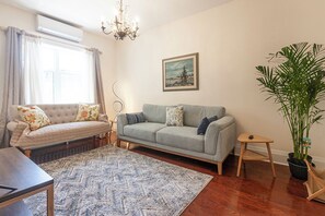 Ein Wohnzimmer mit einer Couch, einem kleinen Tisch, einer Topfpflanze, einem Gemälde an der Wand und einer Pendelleuchte.