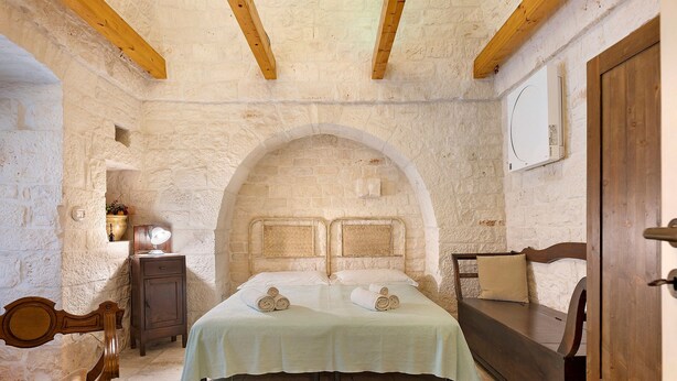 Una habitación con pared de piedra, vigas de madera, una cama con colcha verde, un banco de madera, una mesita de noche con lámpara y un armario de madera.