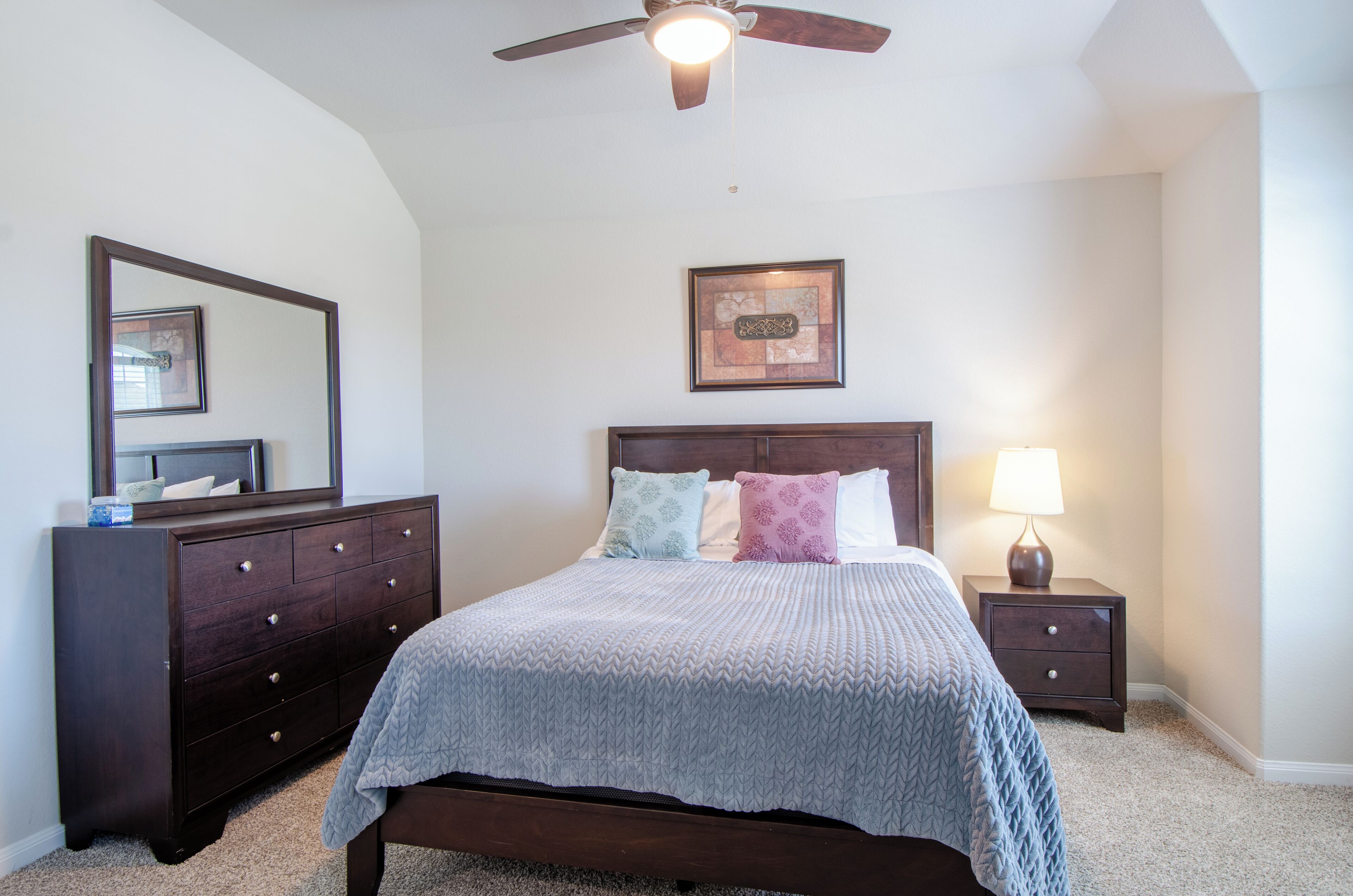 A bedroom with a bed, dresser, nightstand, lamp, ceiling fan, and a framed picture on the wall.