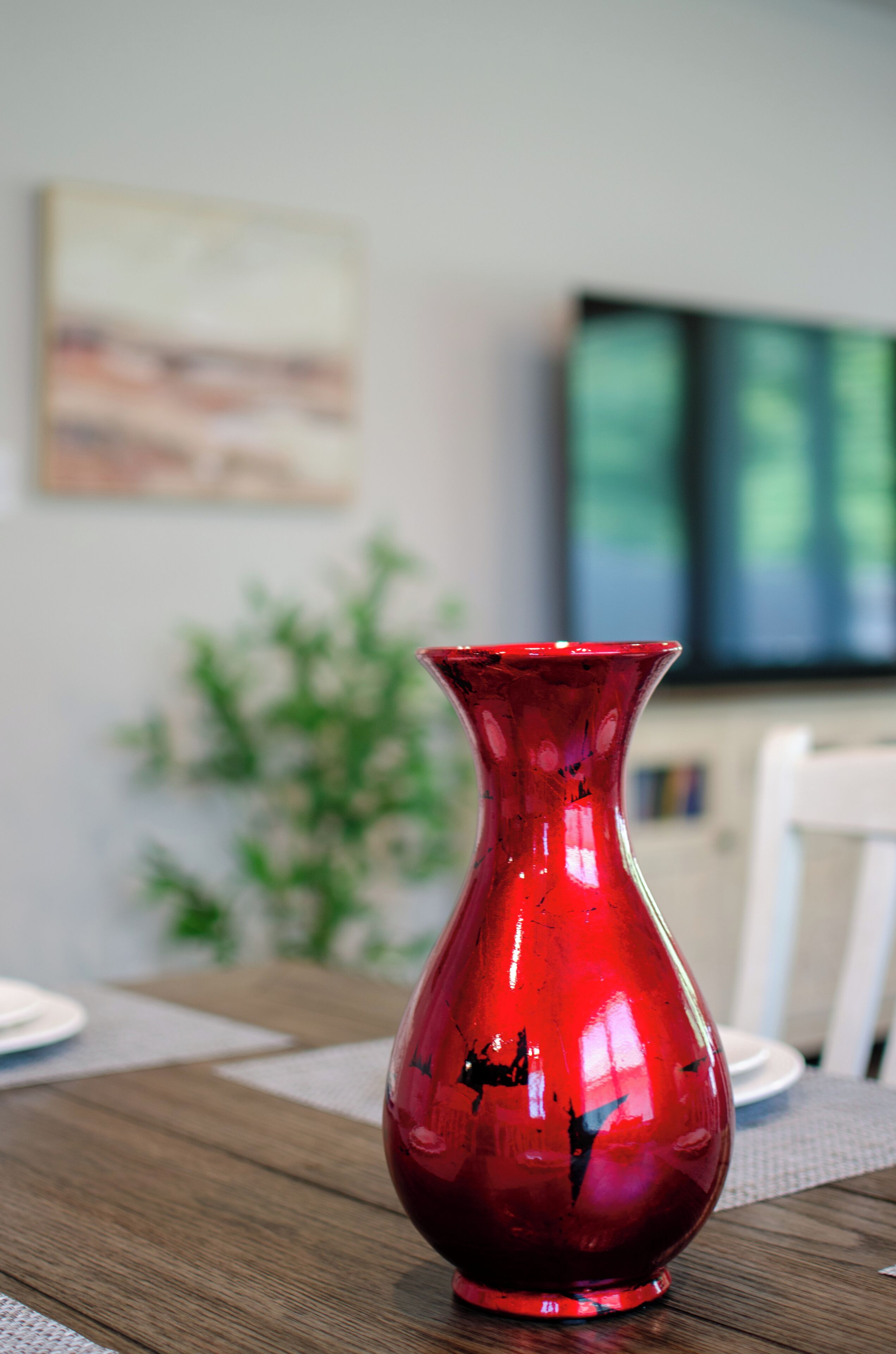 A red vase on a wooden table with a painting and a plant in the background.