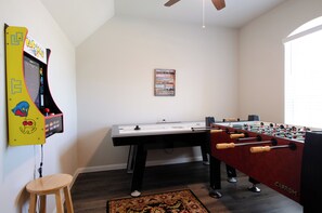 Una habitación con una mesa de futbolín, mesa de futbolín con superficie roja y patas negras, un taburete de madera pequeño y una máquina de juegos montada en la pared.