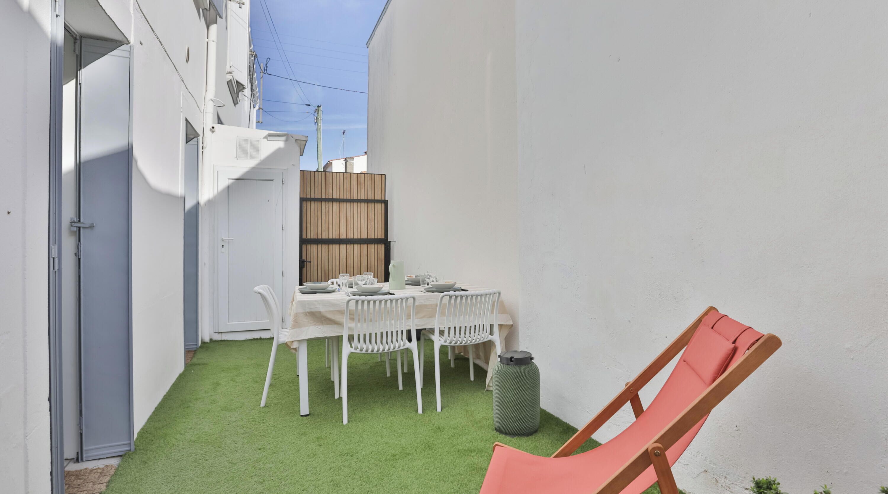 Une petite terrasse avec une table dressée pour deux personnes, des chaises blanches, une poubelle verte et un fauteuil de jardin rouge.