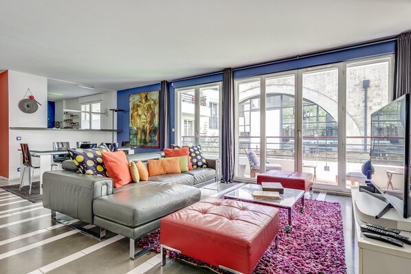 Ein modernes Wohnzimmer mit Sofa, Hocker und Couchtisch, großen Fenstern und Blick ins Freie.