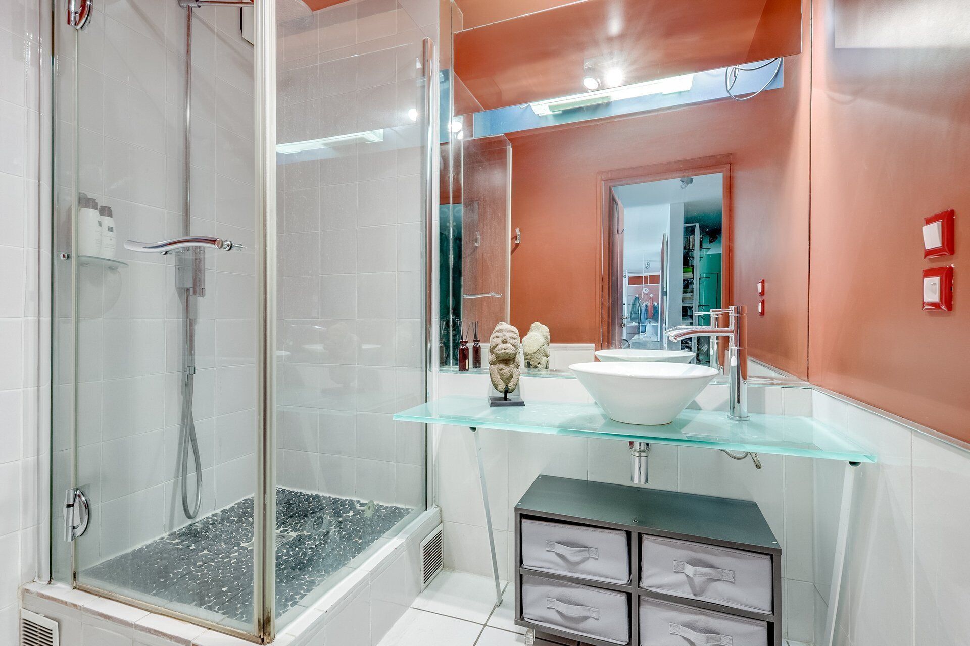 Un baño moderno con cabina de ducha de vidrio, un lavabo blanco y un estante de vidrio con adornos.