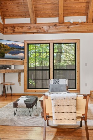 Ein Zimmer mit Holzboden, einem großen Fenster mit Blick auf Bäume, einem Fernseher auf einer Holzablage und einem Sessel mit gemustertem Kissen.