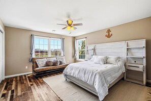 Ein Schlafzimmer mit einem Bett, Deckenventilator, Fenster mit Vorhängen, Holzboden und weißem Kopfteil.