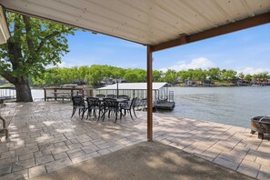 A covered patio with a table and chairs, a grill, and a view of a lake with houses in the background. - Lakefront Home With Stunning Lake Views (Sunrise Beach)